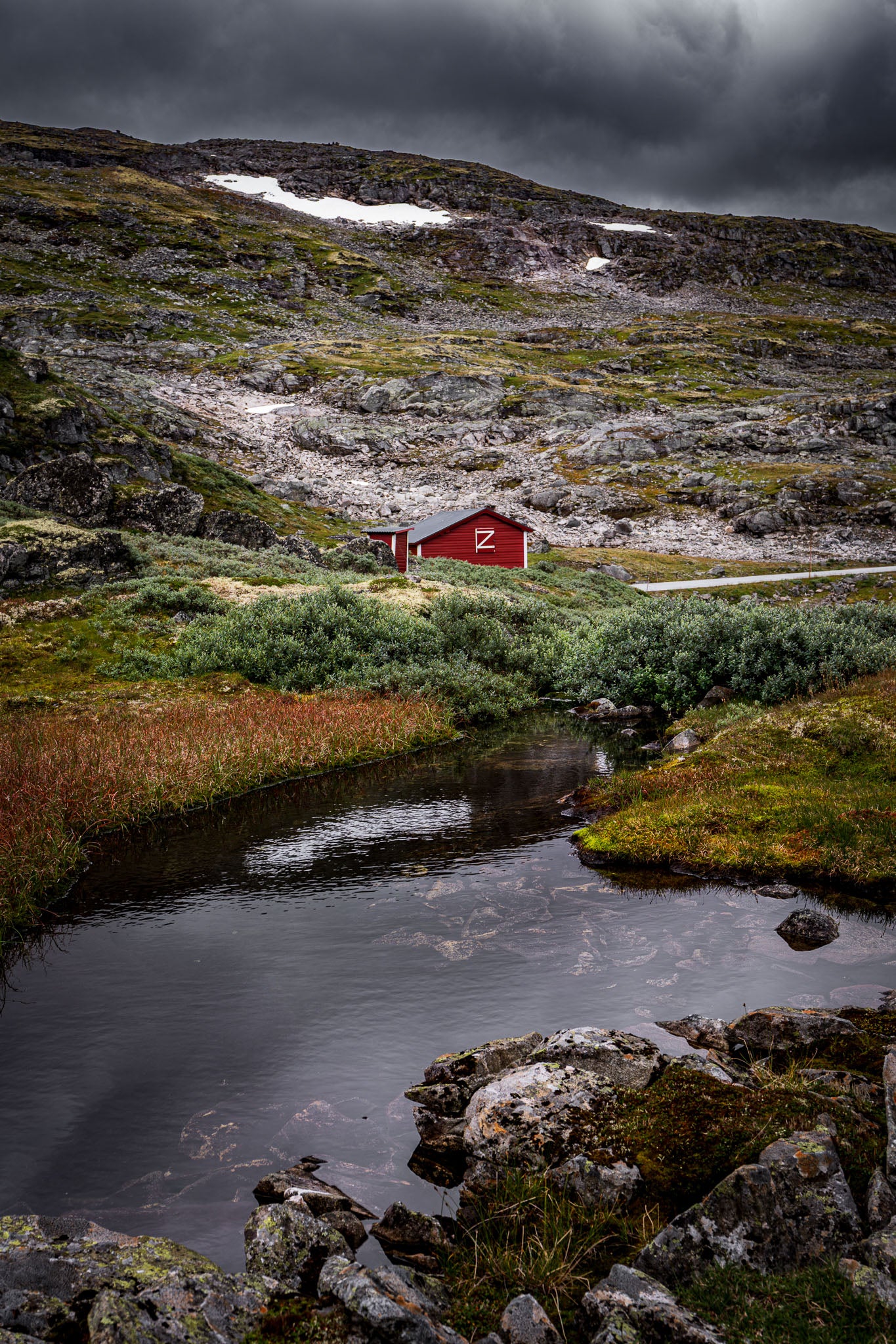 Fine art photograph of a red cabin in Jotunheimen National Park, Norway, set against a backdrop of rocky mountains and moody skies.